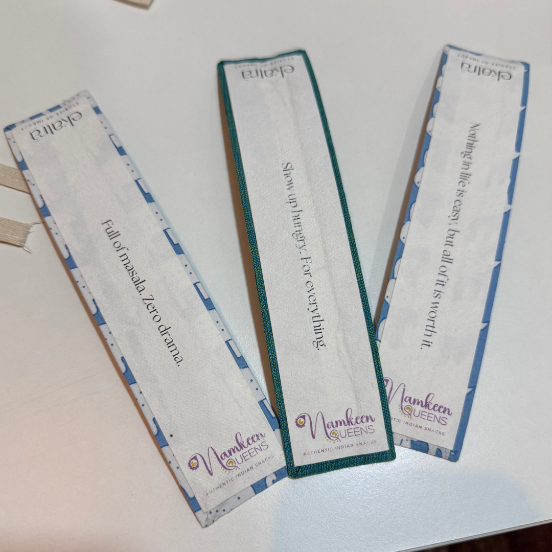 Three long rectangular printed cards or labels laid on a white tabletop. Each card has a decorative border and printed text, including the visible brand name "Namkeen Queens" and "Ekatra" and phrases on each one as follows : Full or masala. Zero drama.
Show up hungry. For everything. 
Nothing in life is easy, but all of it is worth it. 