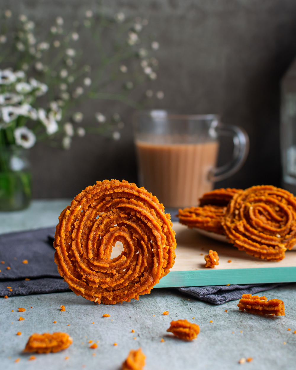 A whole Chakli in focus with Bite-sized crispy snacks arranged on a flat surface with a cup of tea in the background and decorative flowers to the side.